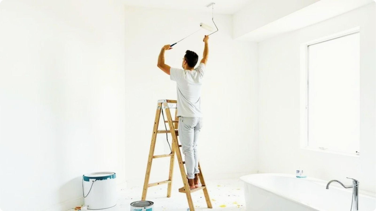 Man on ladder rolling white paint on bathroom ceiling, showing DIY paint application.