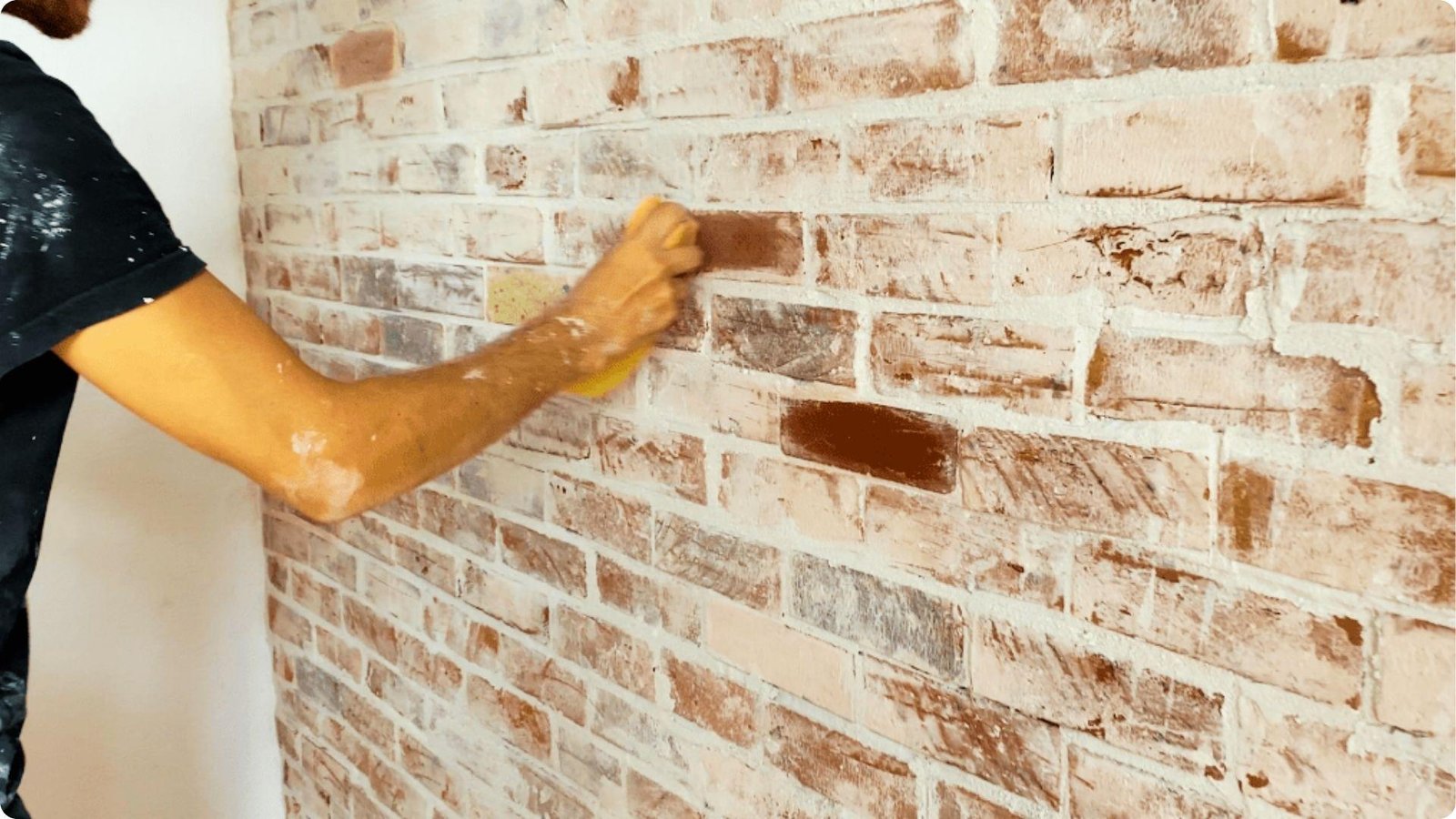 A person uses a sponge to dab whitewash onto an interior brick wall.