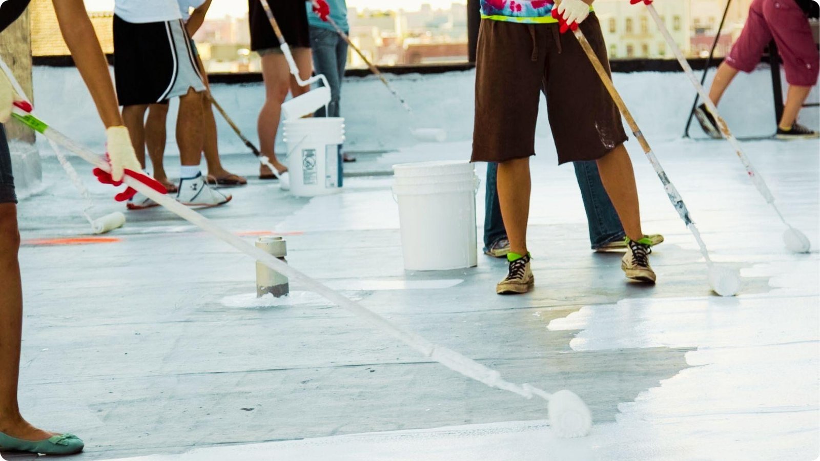Group of people in casual clothes roll white coating onto a flat rooftop with buckets and paint rollers.