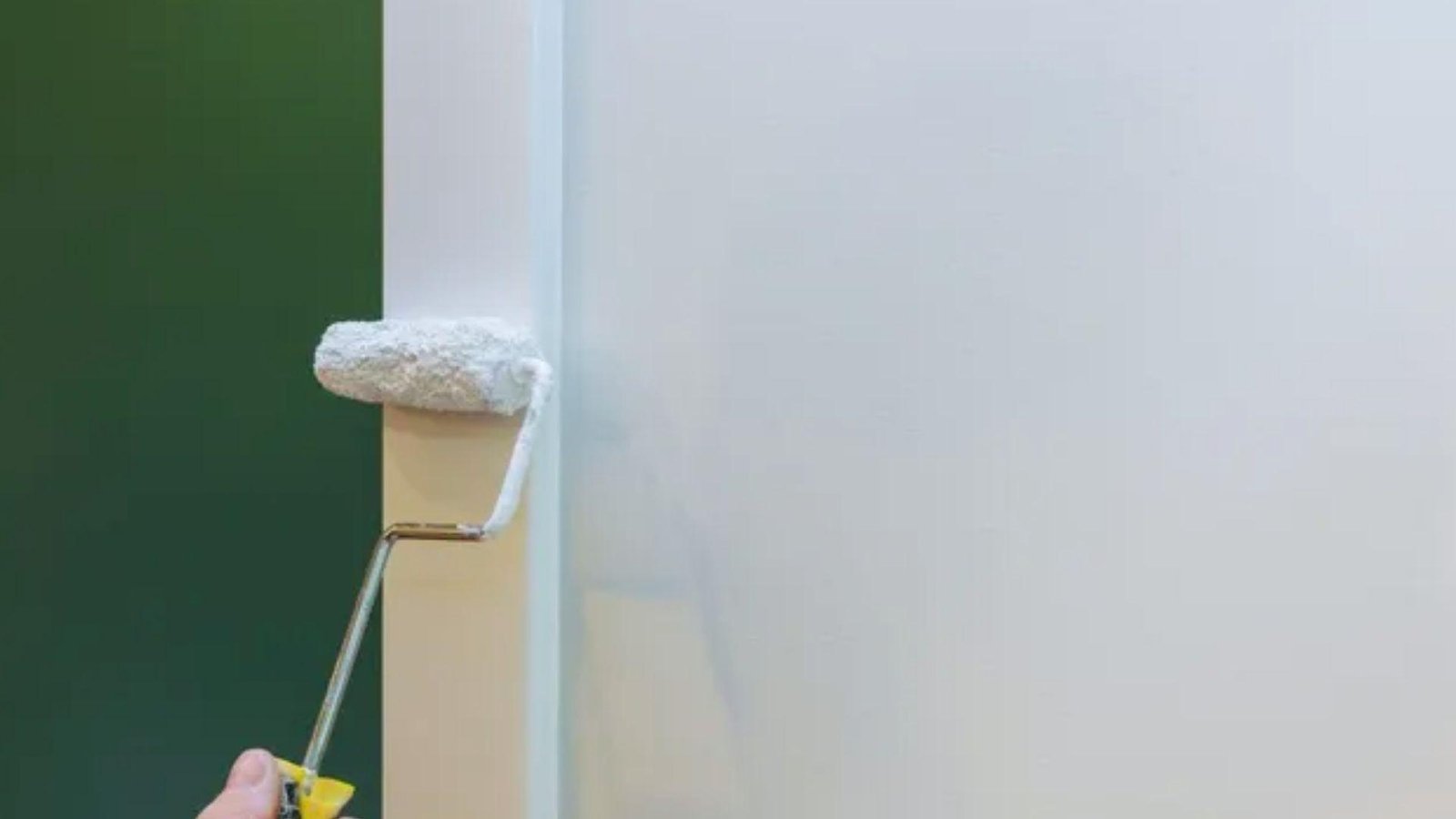 Person using small paint roller to apply white trim paint on interior door casing against dark green walls