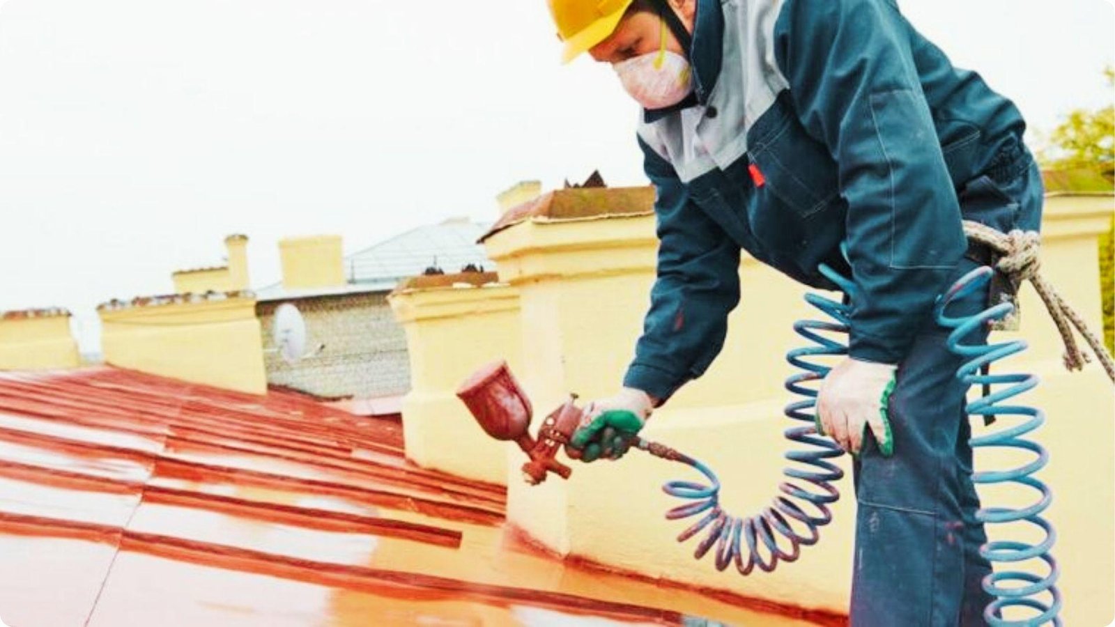 Roofing contractor in a hard hat and respirator uses a spray gun to apply red coating on a sloped roof.